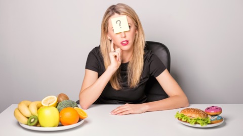 une femme assise à table entre deux assiettes contenant des aliments très différents s'interroge sur le bon régime à suivre